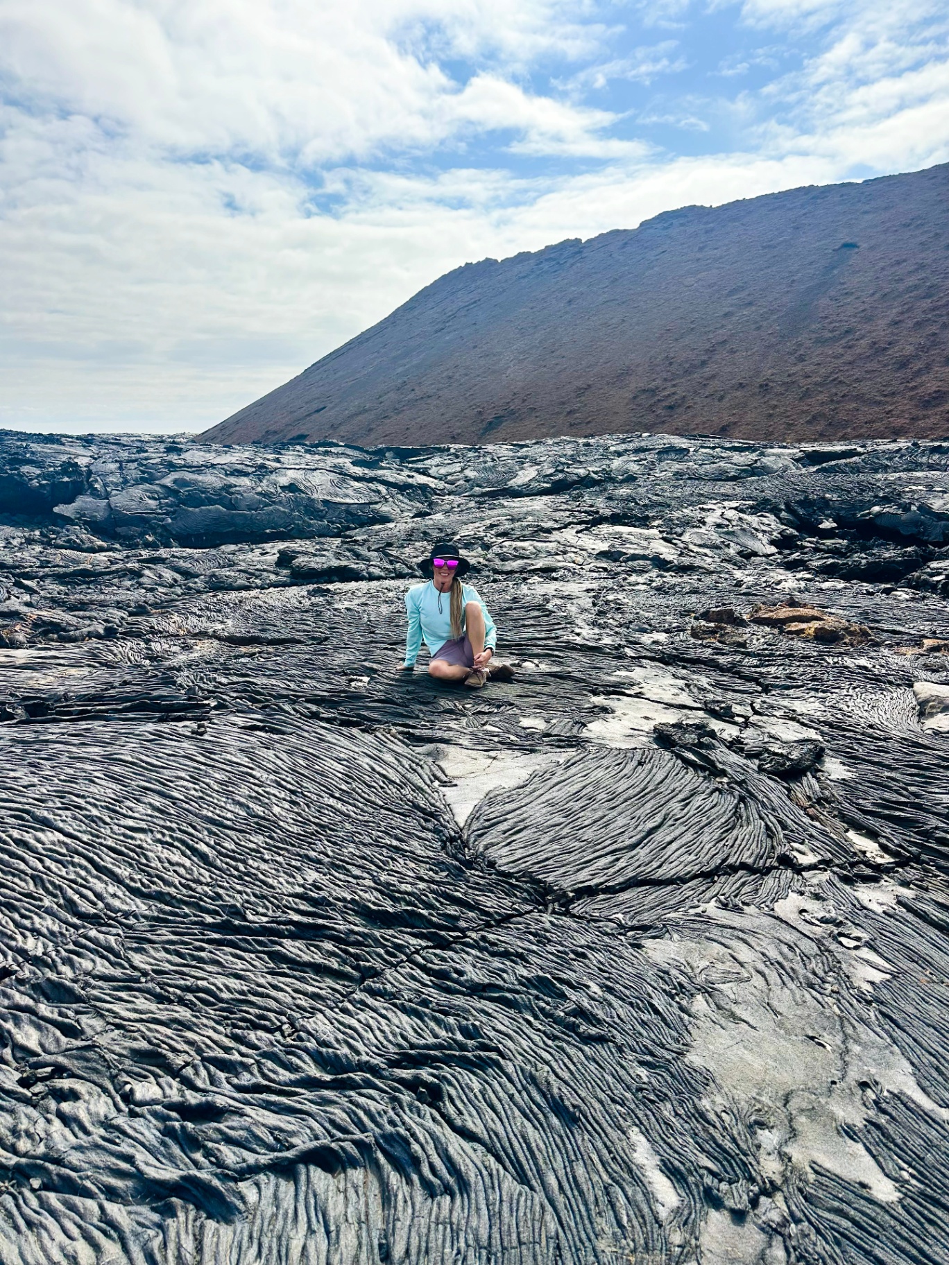 Nicci Grotefendt sits on a hardened lava field in the Galapagos Islands during an expedition landing, a volcanic cone rising behind her.